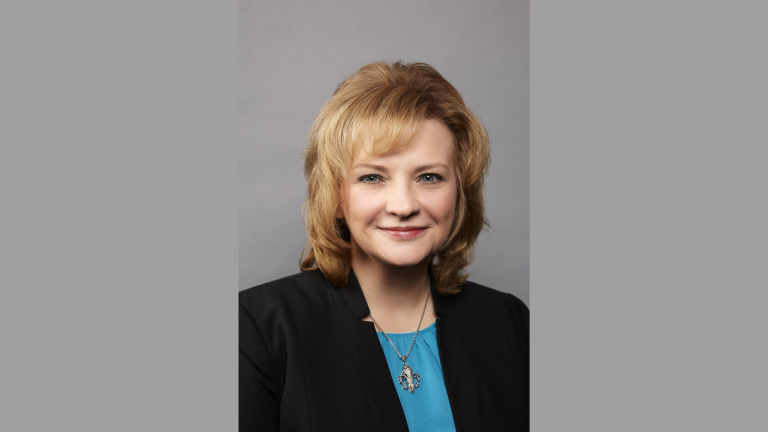 Portrait of Sarah Powell, newly appointed General Counsel of Planet Fitness, wearing a black blazer and blue blouse against a gray background.