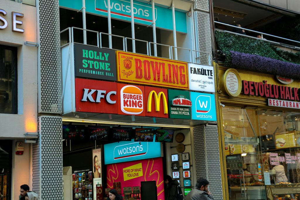 Exterior view of several global franchise storefronts, including KFC, Burger King, McDonald’s, Papa John’s, and Watsons, above a crowded sidewalk in a busy shopping district.