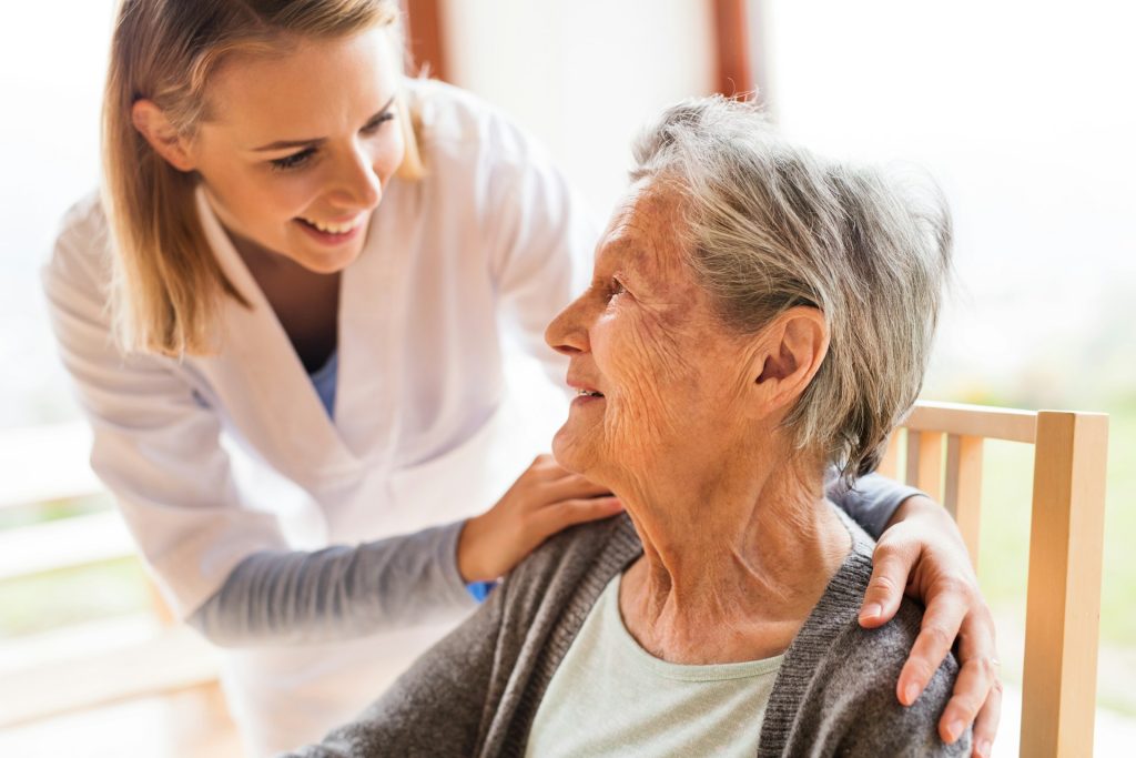 Caregiver smiling and offering support to an elderly woman sitting in a chair during a home care visit.