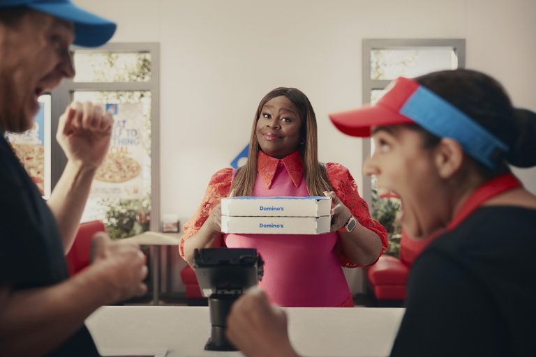 Retta holds Domino’s pizza boxes while employees cheer in a Domino’s store.