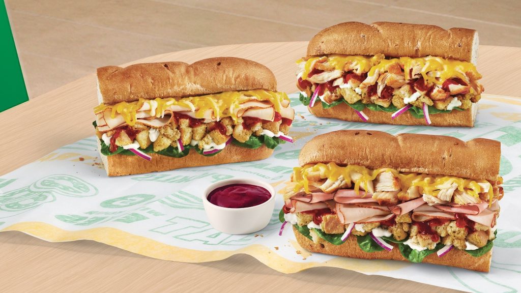 Three Subway holiday subs topped with cheese, stuffing, cranberry sauce, onions, and greens on a table