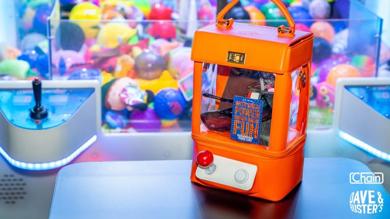 Bright orange claw-machine purse with clear panels sits by arcade claw controls and colorful plush prizes