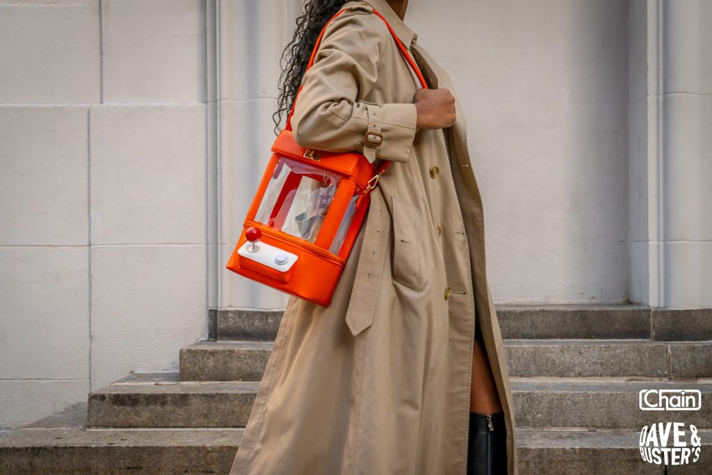 Person in a trench coat carries bright orange claw-machine purse with clear sides on city steps