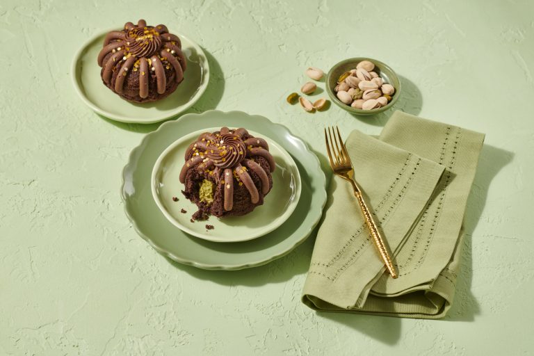 Chocolate Nothing Bundt Cakes Bundtlets topped with frosting and gold sprinkles, styled with pistachios