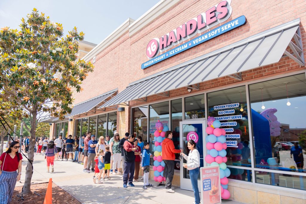 Crowd of customers lining up outside a 16 Handles store on grand opening day decorated with pink and blue balloons