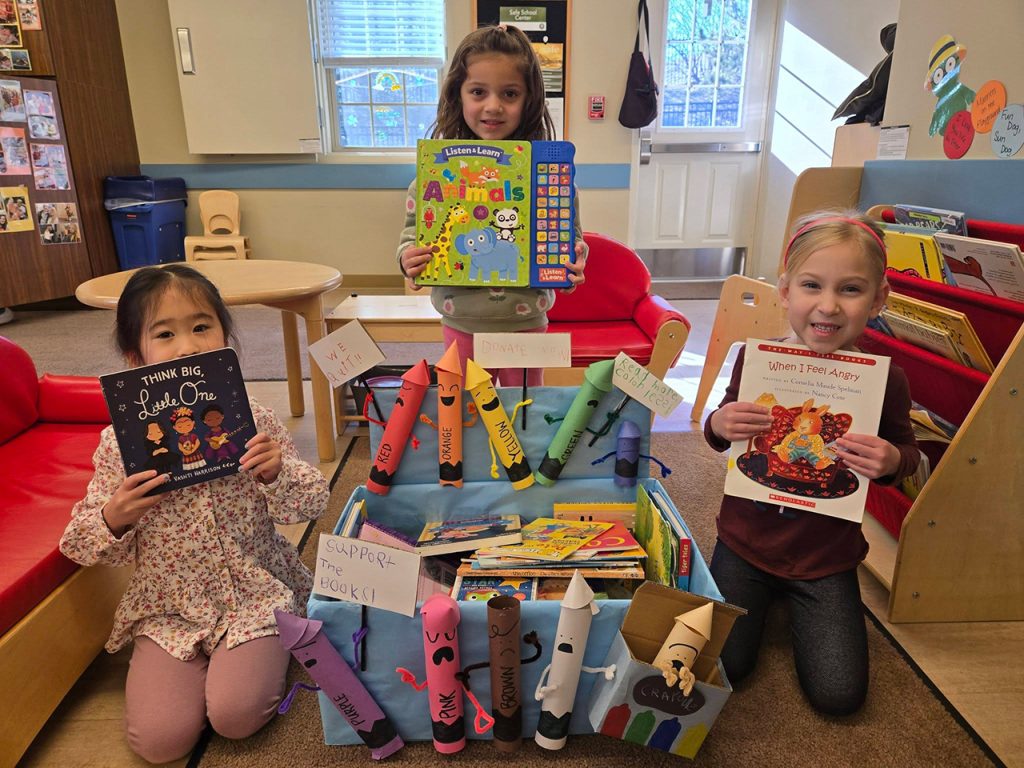 Three children hold books beside a donation bin in a Primrose classroom during a book drive