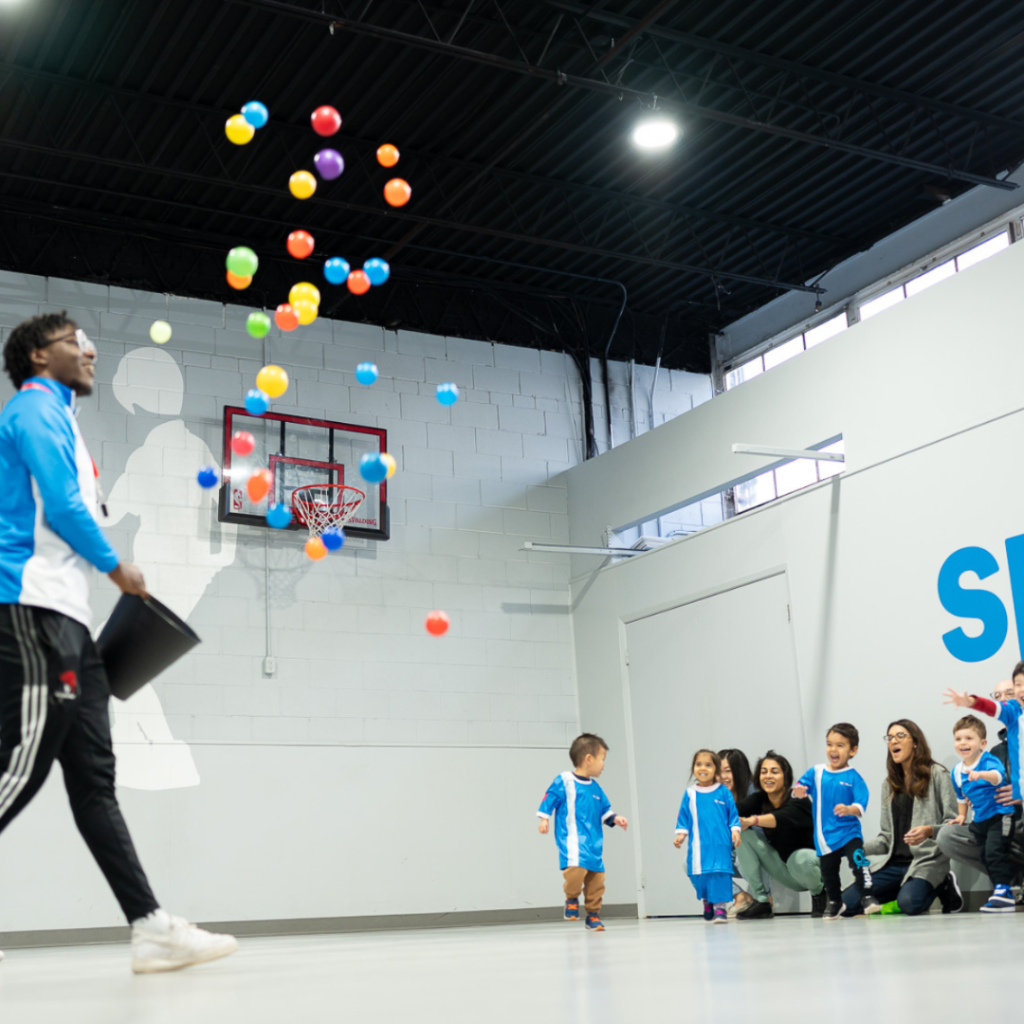 Coach tosses colorful balls into the air while children in Sportball uniforms react during an indoor class activity.