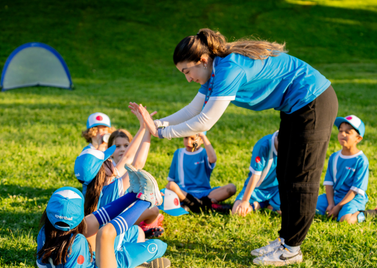 Sportball Turns Early Movement Into a Lifelong Advantage Sportball coach high-fives a child during an outdoor class while other children in blue uniforms sit on the grass nearby.