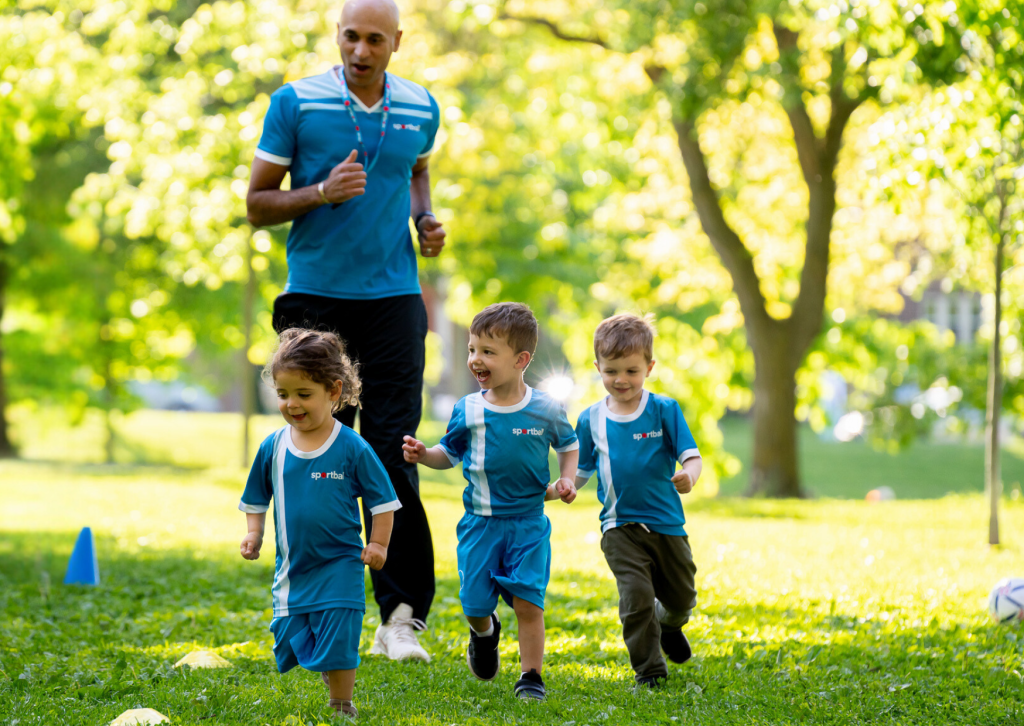 Sportball coach runs with young children in blue uniforms during an outdoor class on the grass.