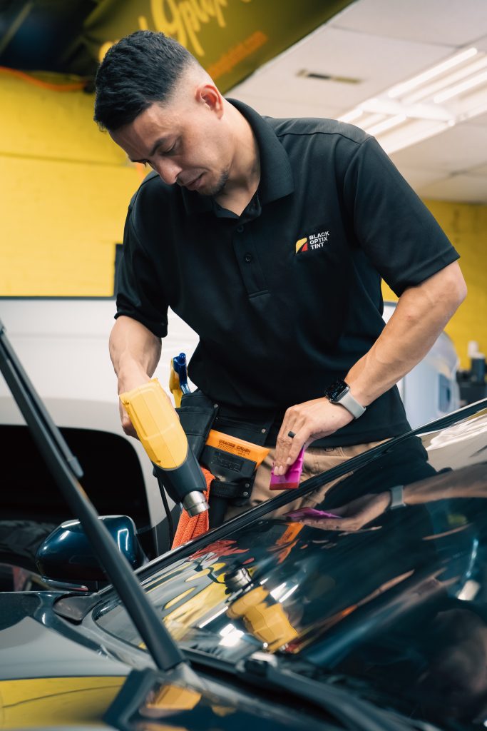 Black Optix Tint technician using a heat gun and squeegee to apply window tint film to a vehicle windshield