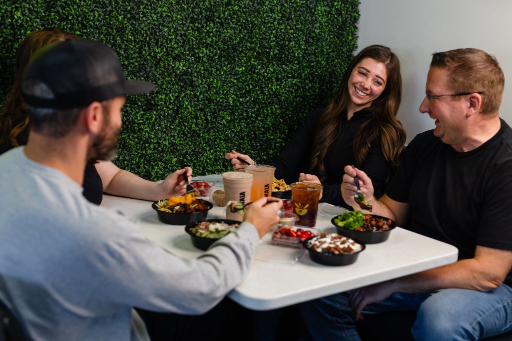 People laughing and sharing Konala protein bowls and beverages at a table inside a Konala location