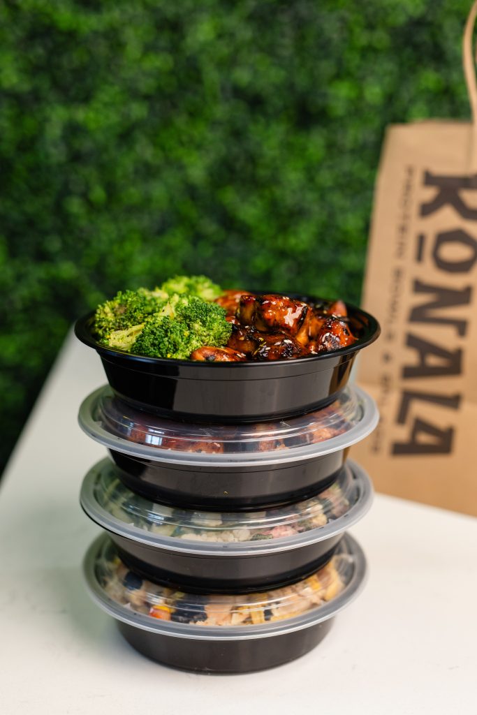 Stack of four Konala protein bowls with a teriyaki chicken and broccoli bowl on top next to a Konala branded bag