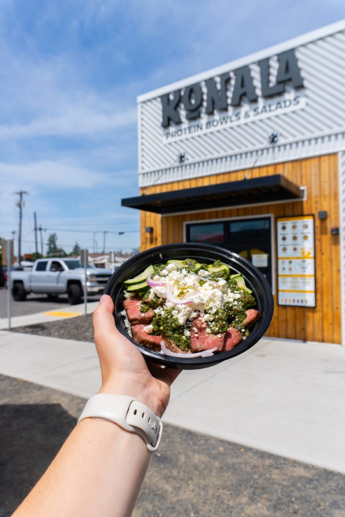 Hand holding a Konala Greek steak protein bowl with feta and chimichurri in front of a Konala location