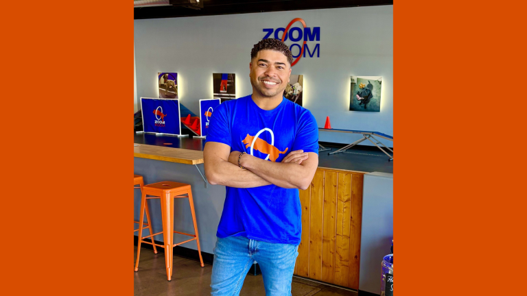 Zoom Room Names Darris Cooper Director of Training and Socialization Darris Cooper standing inside a Zoom Room dog training gym wearing a branded blue shirt