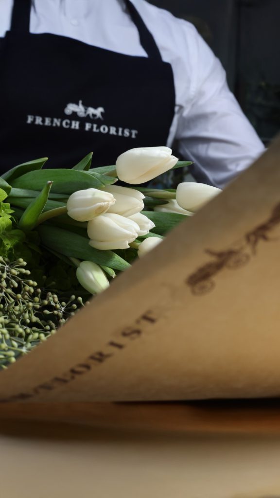 A French Florist team member in a branded apron holding a wrapped bouquet of white tulips and greenery in kraft paper packaging.