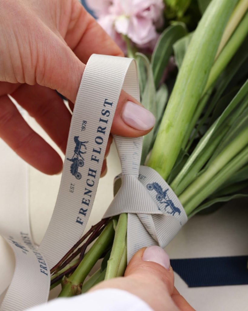 Hands tying a French Florist branded grosgrain ribbon around a fresh bouquet stem, showing the brand's attention to finishing detail.