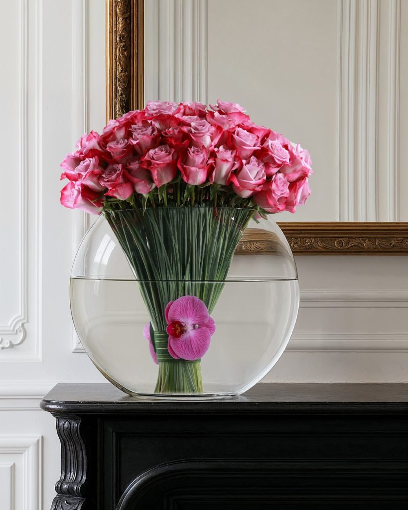 A dramatic French Florist arrangement of pink roses and a floating orchid in a large round glass bowl vase displayed on a dark mantle.