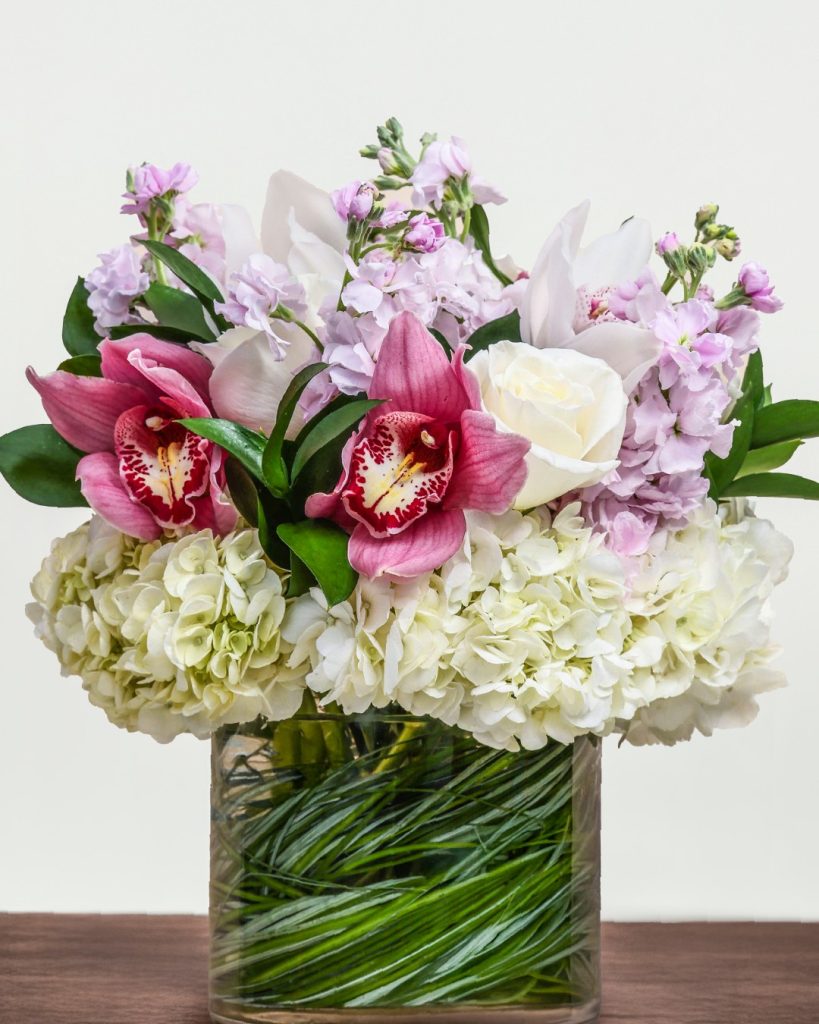A French Florist arrangement featuring white hydrangeas, pink cymbidium orchids, white roses, and lavender stock in a glass vase.