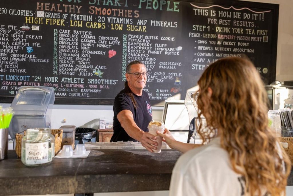 Daniel Boone of HB Protein Smoothies hands a shake to a customer in front of the brand's detailed educational menu board.
