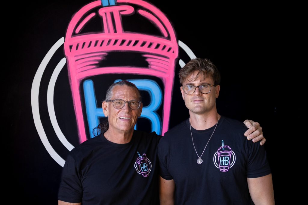 HB Protein Smoothies founders Daniel and Ethan Boone in front of the brand's neon logo sign.
