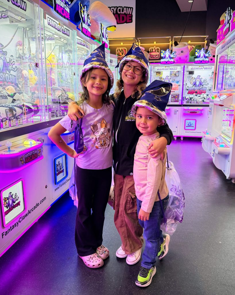 Three kids wearing Fantasy Claw Arcade wizard hats smiling inside the arcade surrounded by claw machines