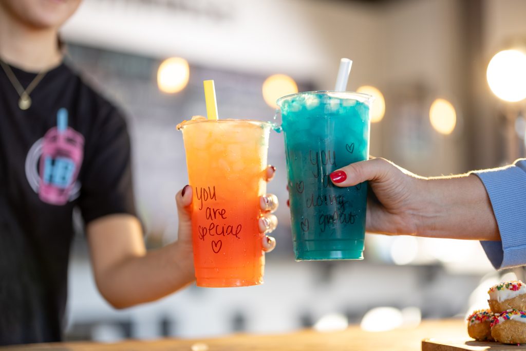 Two hands clink colorful HB Energy drinks in clear cups, one orange reading "you are special" and one teal reading "you are doing great," handwritten in marker with heart doodles.