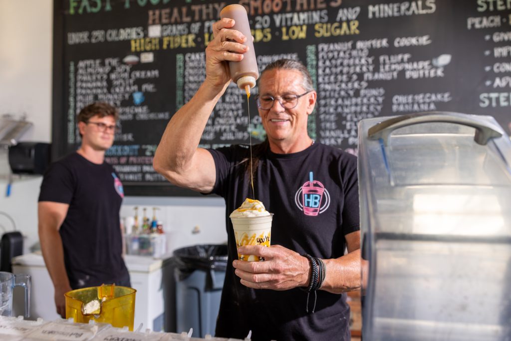 HB Protein Smoothies founder Daniel Boone finishing a shake at the Scottsdale location with the menu board visible behind him.