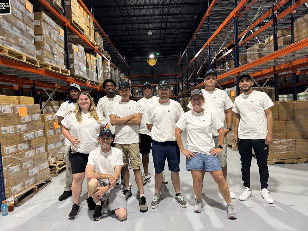 Window Gang of Palm Beach team in matching shirts and Kids Lift caps posing in a warehouse aisle surrounded by donated supplies