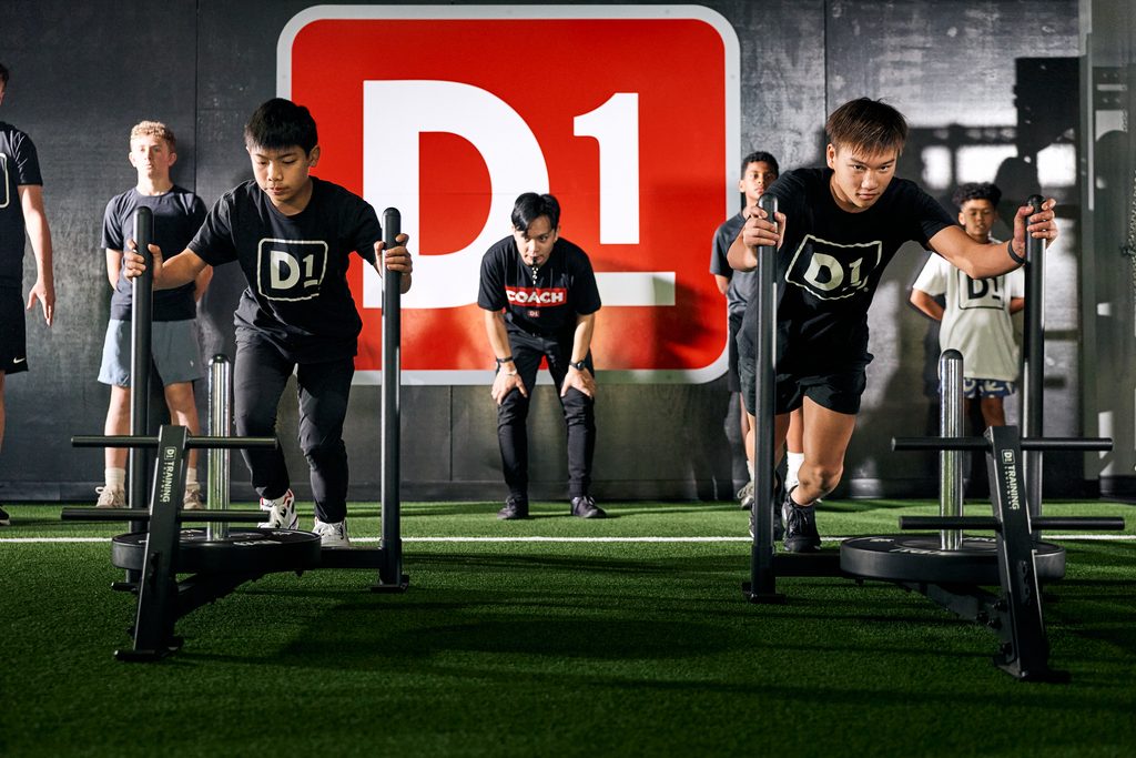 Young athletes performing weighted sled pushes at a D1 Training facility while a coach supervises