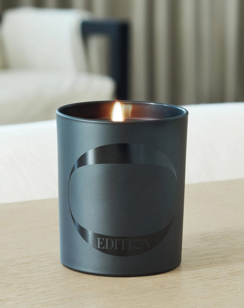 Lit EDITION Hotels candle in a dark glass vessel displayed on a wooden surface in a hotel room.