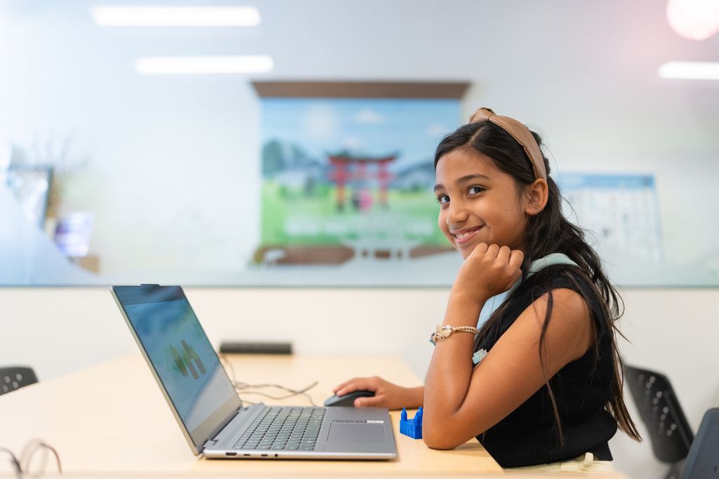 A girl smiles at the camera while working on a laptop at a Code Ninjas dojo table.