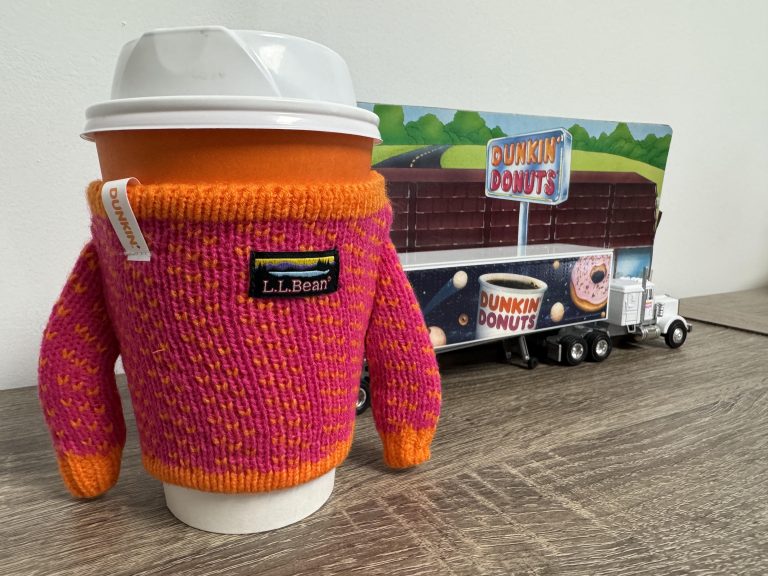 Dunkin’ coffee wearing a pink and orange L.L.Bean sweater beside a Dunkin’ truck collectible