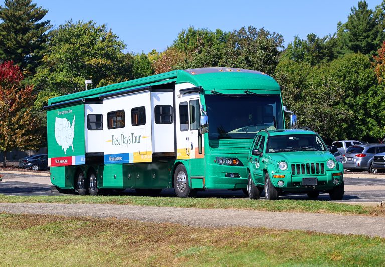 FirstLight Home Care Best Days Tour RV parked beside a green FirstLight-branded Jeep with autumn trees behind